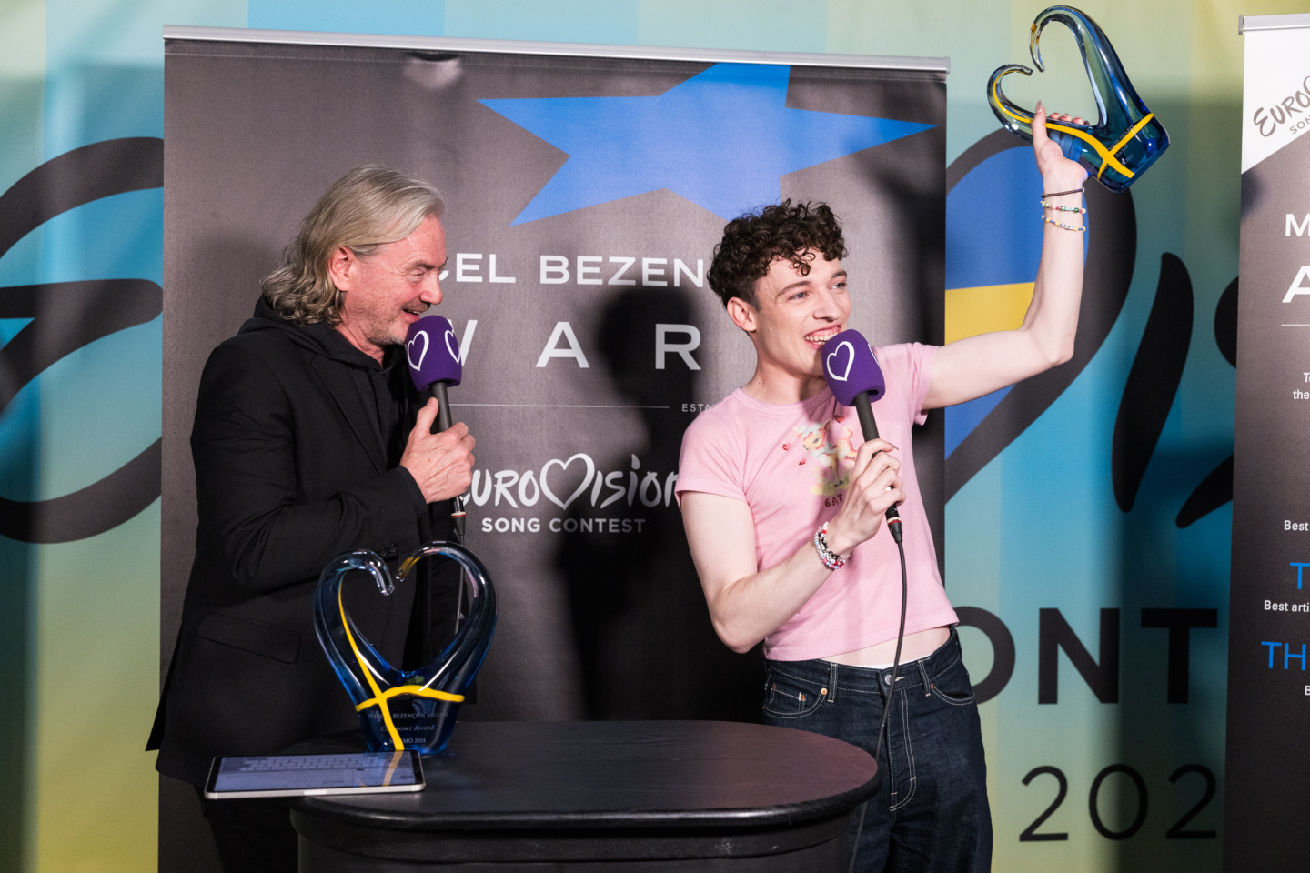 Nemo, dressed in a pink top and black jeans, lifts aloft a blue heart trophy with the swedish flag on it while talking into a eurovision heart branded microphone. Christer Bjorkman looks on and smiles as they do so
