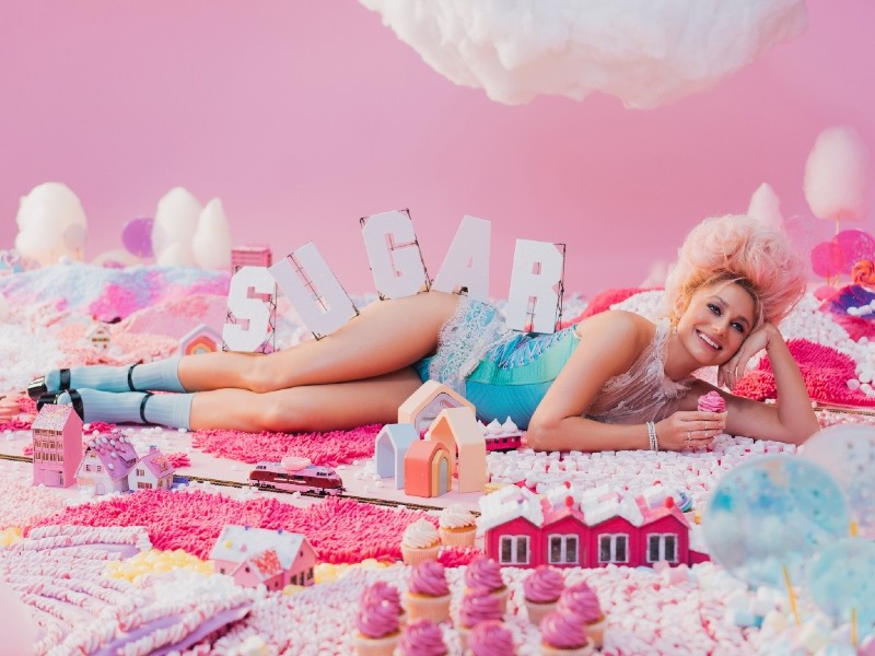 natalia gordienko lying on her side, with the letters of the word SUGAR across her legs and torso. she is surrounded by pink, candy-inspired imagery.
