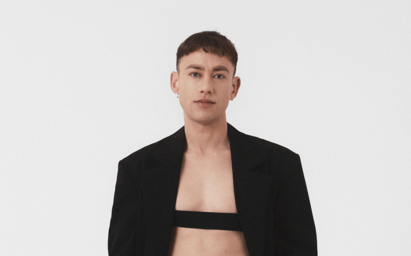 Olly Alexander wearing a black suit with a black bar of fabric across his chest.
