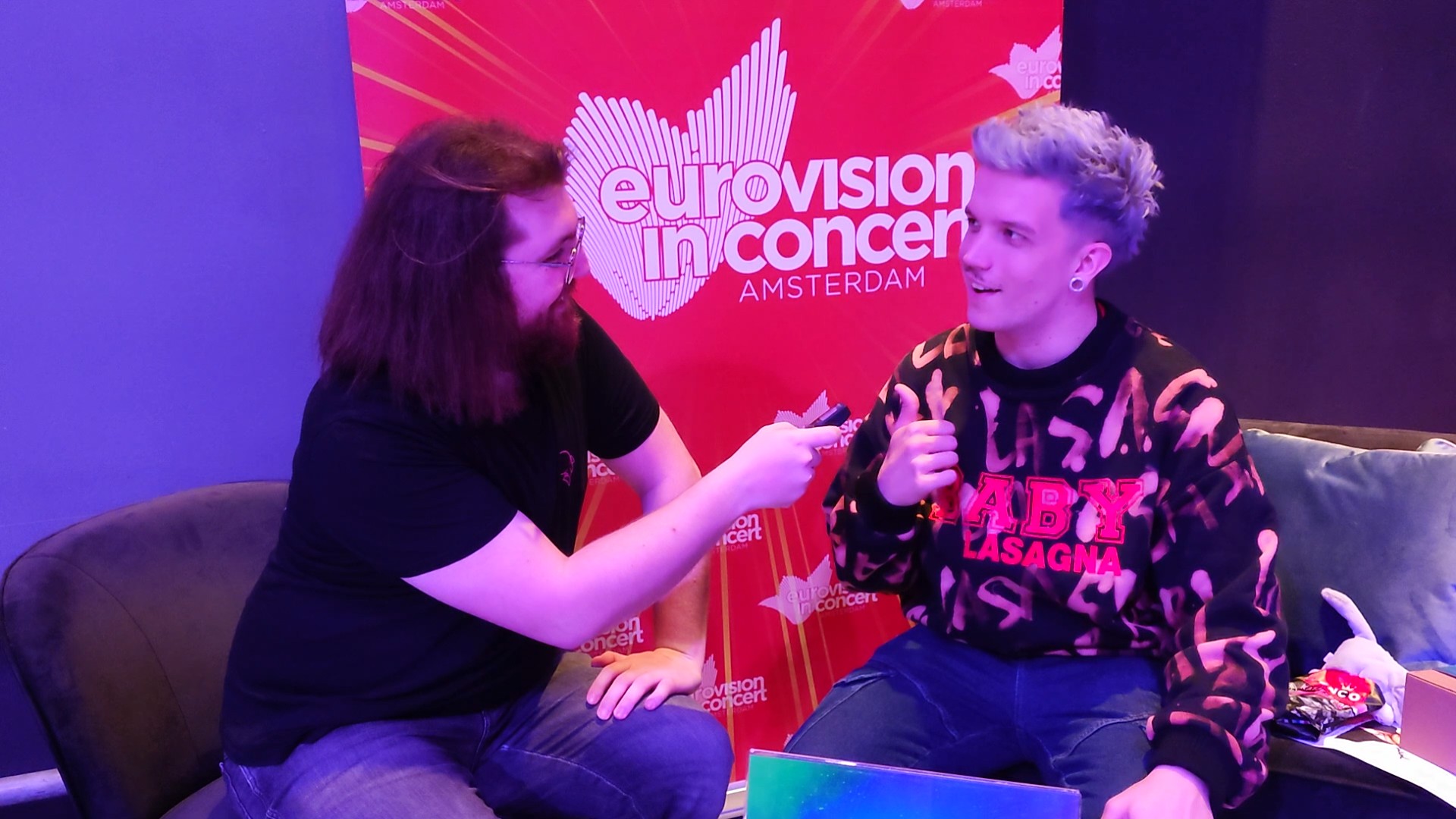 Baby Lasagna and Angus sitting down for an interview with the Eurovision in Concert logo behind them